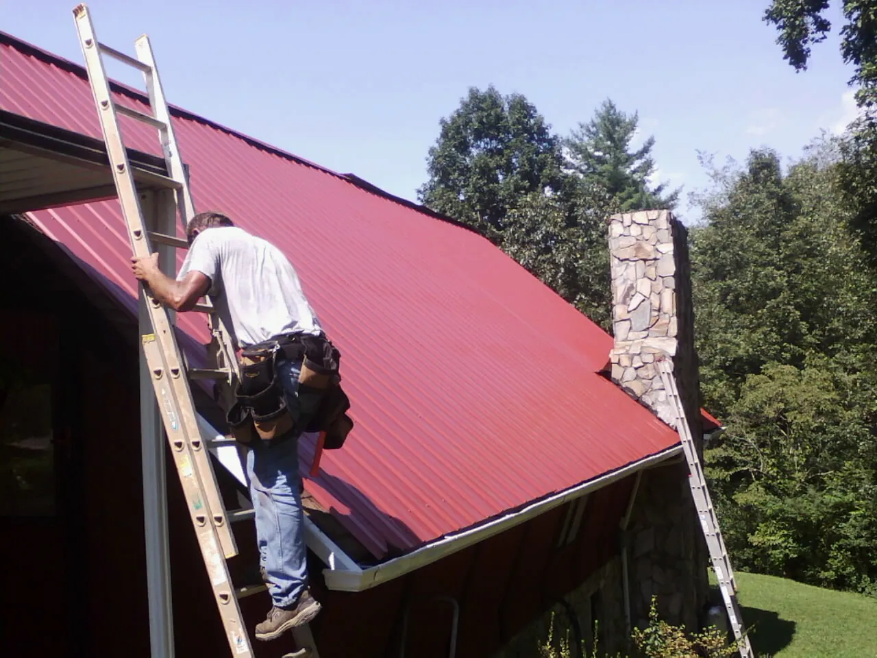 Roofer on a ladder beside a red metal roof and stone chimney — Soffit & Fascia Repair by Roofing Brothers in Bay Village